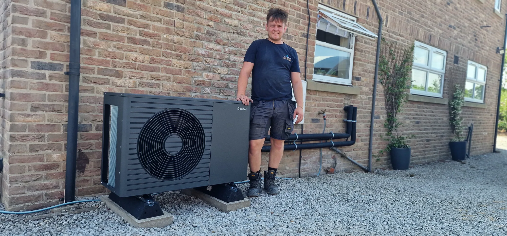 PCR Plumbers engineer with a completed Vaillant heat pump installation PCR Plumbers heating engineer beside a newly installed Vaillant air-source heat pump at a rural brick property near York and Selby